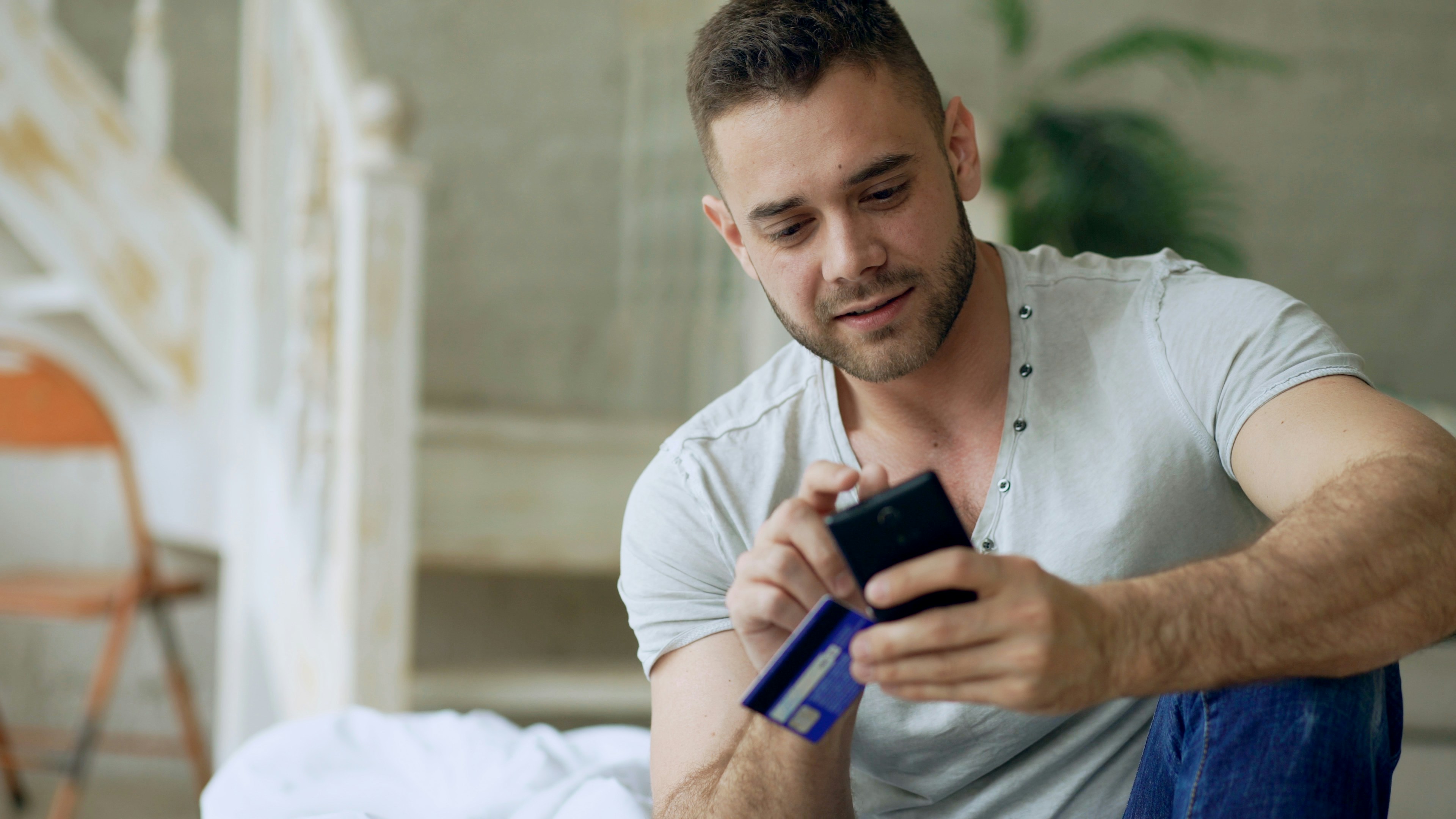 Attractive young man with smartphone and credit card shopping the internet sit on bed at home